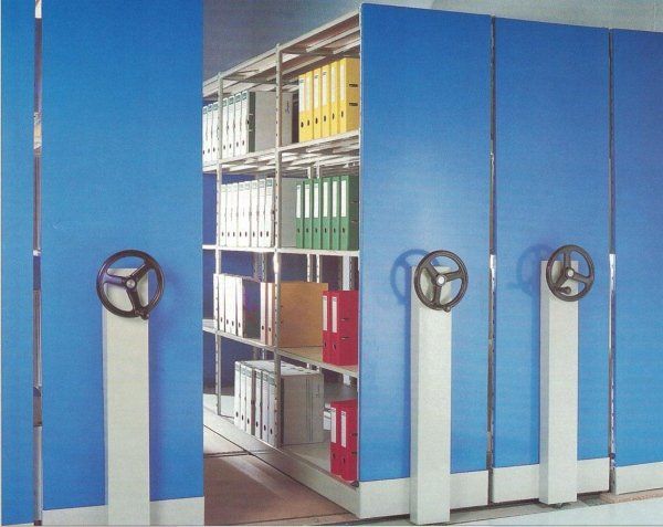 Un sistema di archiviazione mobile con ripiani scorrevoli blu, binari metallici e ruote a manovella per accedere alle file di raccoglitori da ufficio.