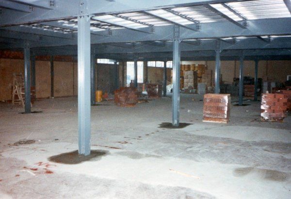 Interno di un magazzino industriale caratterizzato da travi di supporto in acciaio, pavimento in cemento e pallet di materiali impilati.