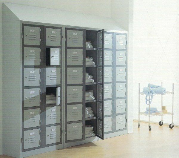 Una parete di armadietti in metallo grigio per la biancheria o gli asciugamani in una stanza luminosa, con un carrello su ruote contenente lenzuola piegate nelle vicinanze.