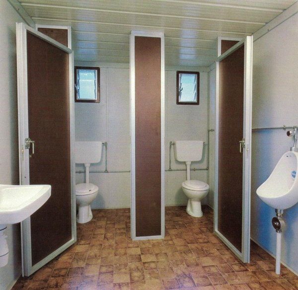 Interno di un bagno pubblico con due cabine WC private dotate di porte marroni, un divisorio centrale e un orinatoio bianco.