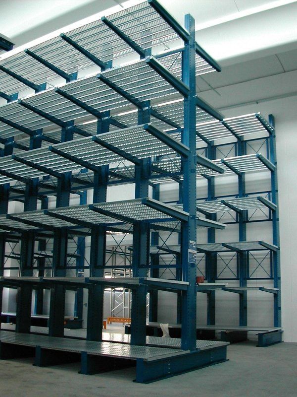 All'interno di un magazzino, si trova un alto sistema di scaffalature industriali a sbalzo di colore blu, con ripiani in grigliato metallico.
