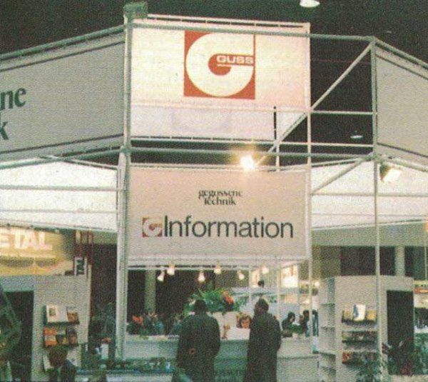 Uno stand fieristico con l'insegna di Guss and Johnson Kjekshus, dove i visitatori sono in attesa presso un punto informazioni.