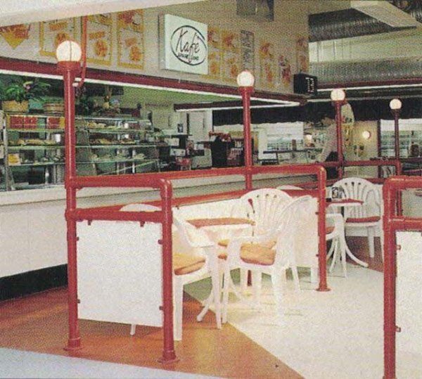 Una bancarella di cibo con tavoli e sedie bianchi, delimitata da ringhiere metalliche rosse, con una piccola insegna con la scritta 