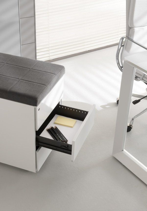 Un piedistallo mobile bianco con sedile imbottito grigio e cassetto aperto contenente articoli di cancelleria, posizionato vicino a una scrivania.