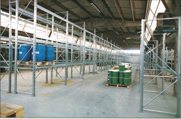 Magazzino industriale con scaffalature metalliche vuote e pallet di fusti chimici blu e verdi sul pavimento di cemento.