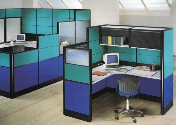 Due postazioni di lavoro con pannelli color verde acqua e blu, scrivanie, sedie e computer su un pavimento di colore chiaro.