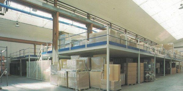 Interno di un magazzino caratterizzato da un soppalco metallico con scatole impilate sopra unità di stoccaggio pallettizzate a livello del suolo.