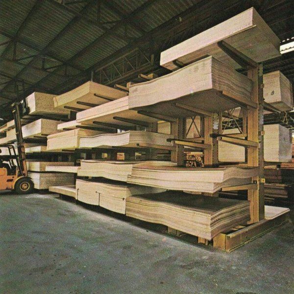 In un magazzino, un carrello elevatore è posizionato accanto a scaffalature a sbalzo alte, su cui sono impilati numerosi pannelli di compensato di grandi dimensioni.