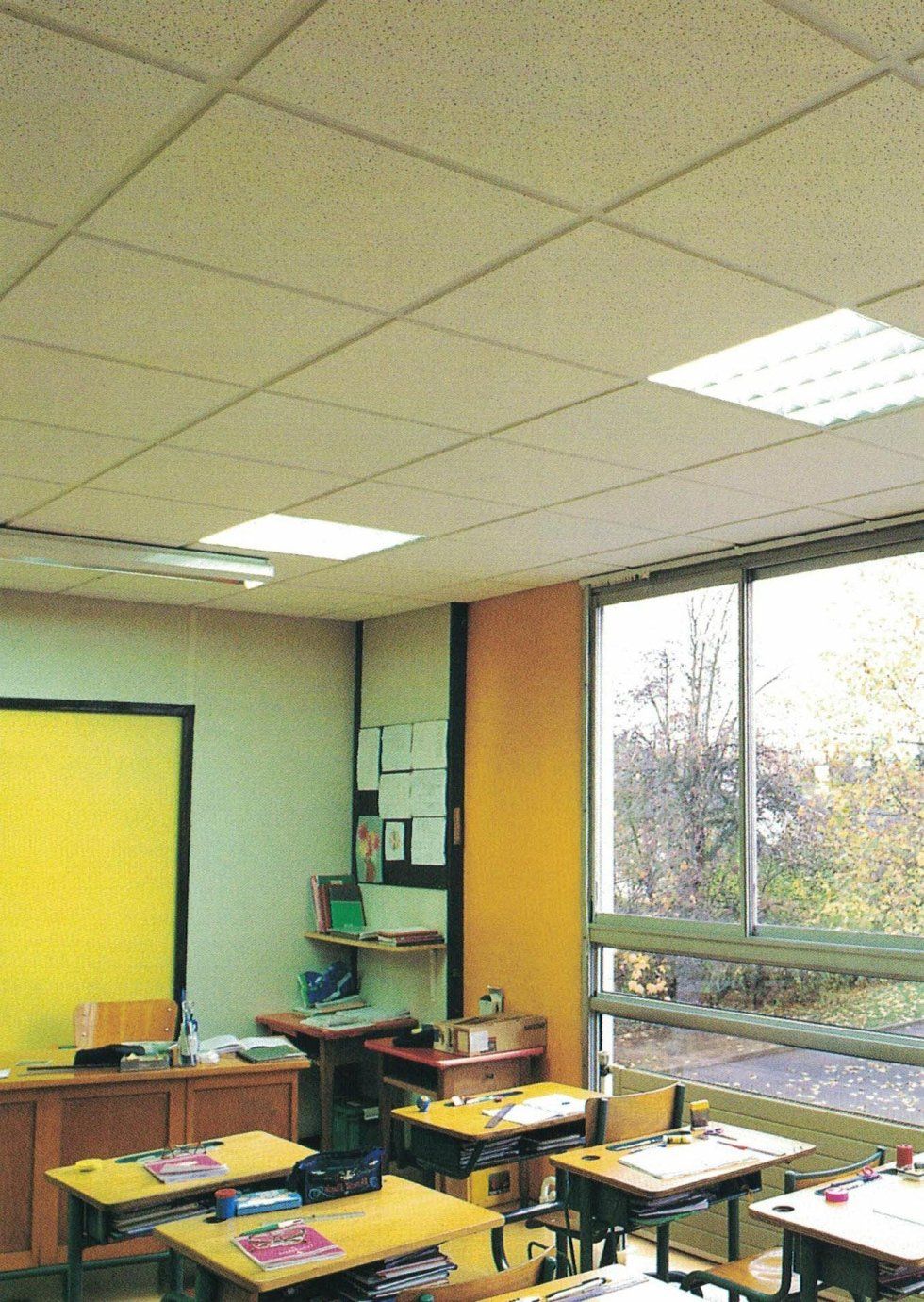 Un'aula con file di banchi di legno, una grande finestra che si affaccia sugli alberi, una parete gialla e un controsoffitto con luci.