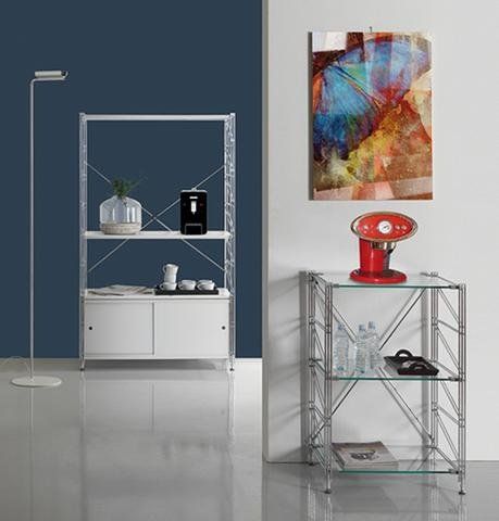 Una scaffalatura in metallo e un espositore con piano in vetro, addossati a una parete blu scuro e bianca, con un frullatore rosso e delle opere d'arte.