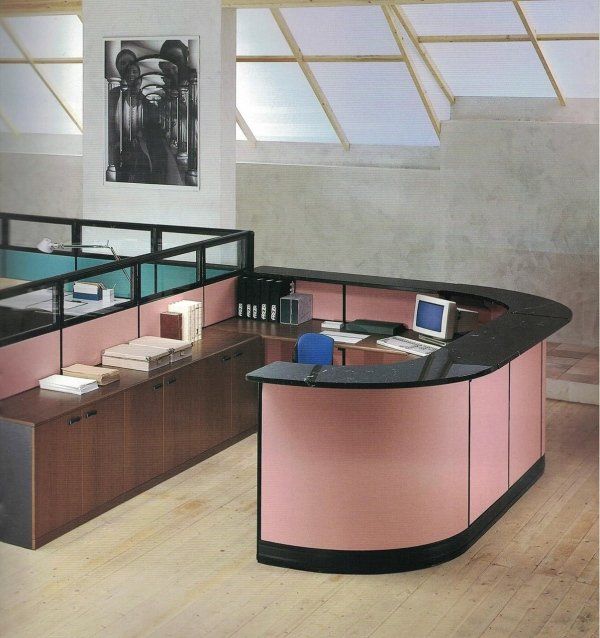 Un moderno bancone reception per ufficio con frontale curvo rosa e piano di lavoro nero, posizionato in un ambiente luminoso con pavimento in legno.