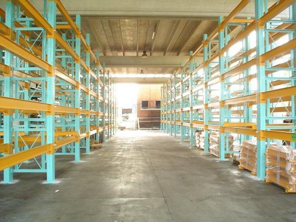 Un corridoio tra due file di scaffalature industriali alte e azzurre, con travi metalliche gialle, all'interno di un magazzino.