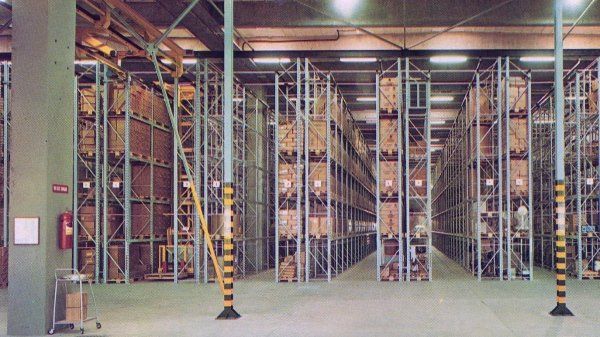 In un grande magazzino ben illuminato, delimitato da pali di sicurezza gialli e neri, file di scaffalature industriali piene di scatole.