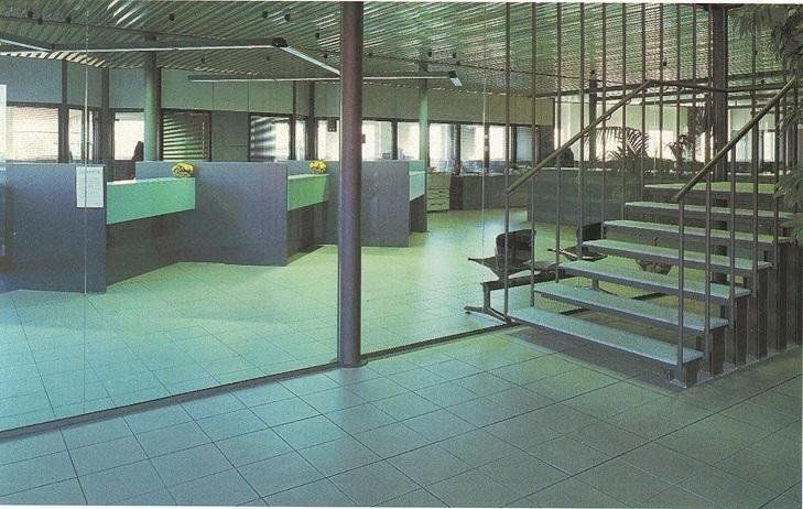 Vista interna di un moderno ufficio vuoto con postazioni di lavoro verde chiaro, pavimenti in piastrelle di colore chiaro e scale.
