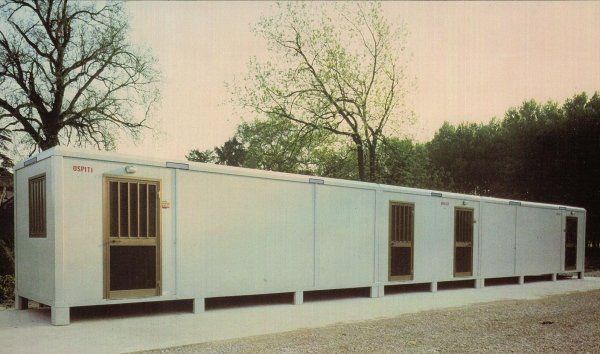 Un lungo edificio modulare bianco e rettangolare, con diverse porte e finestre, sorge su una superficie di ghiaia vicino agli alberi.