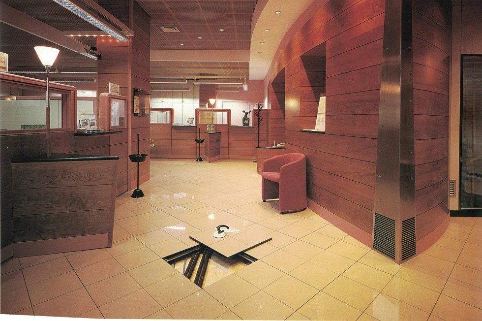 Una moderna hall d'ufficio con pareti in legno color marrone rossastro, pavimento piastrellato beige, una sola sedia e un pannello di accesso a pavimento aperto.