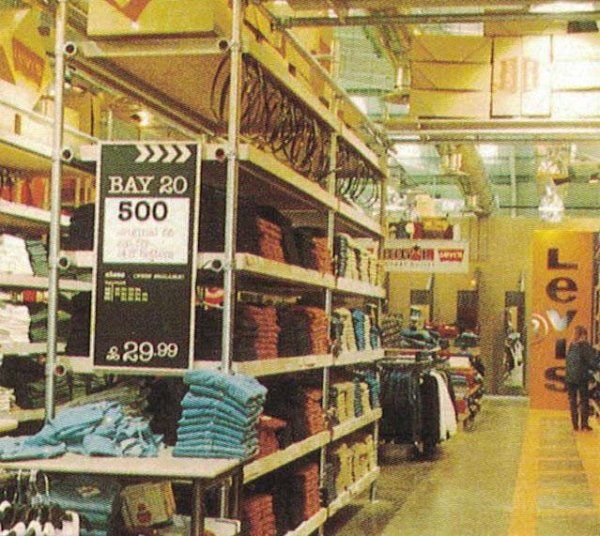 Una corsia di un negozio al dettaglio con scaffali metallici pieni di jeans piegati, un cartello con la scritta 