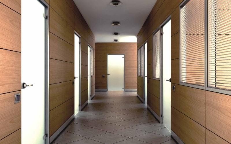 Un corridoio per uffici moderno e luminoso, con pareti rivestite in legno chiaro, pavimenti piastrellati e porte a vetri su entrambi i lati.
