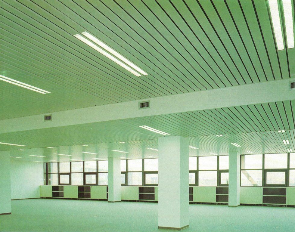 Un ampio ufficio vuoto con soffitto metallico bianco a linee, illuminazione a incasso, colonne portanti e pareti vetrate.