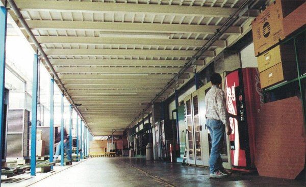 Una persona si trova davanti a un distributore automatico di Coca-Cola sotto una tettoia esterna, con delle scatole impilate sulla destra.