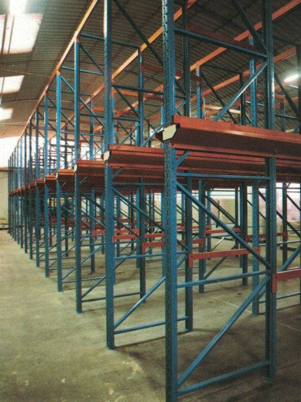 Scaffalature industriali vuote in acciaio per magazzino, con montanti verticali blu e travi orizzontali arancioni, su un pavimento in cemento.