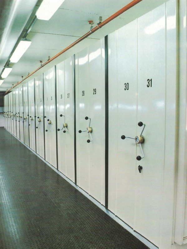 Una fila di scaffalature mobili bianche ad alta densità, con meccanismi manuali a manovella, numerate in sequenza.