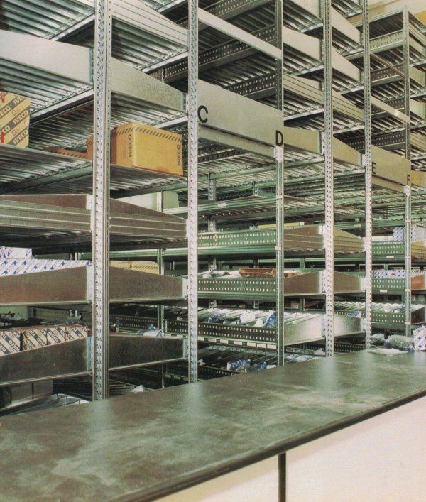 Un'area di stoccaggio con file di scaffalature industriali in metallo, alcune etichettate con lettere, contenenti scatole e oggetti di vario genere.