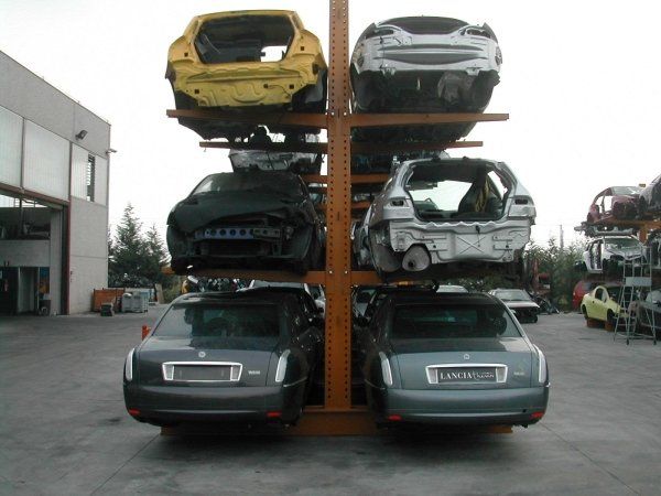 Una struttura metallica industriale in un deposito di rottami, che ospita all'aperto carrozzerie e telai di automobili impilati.