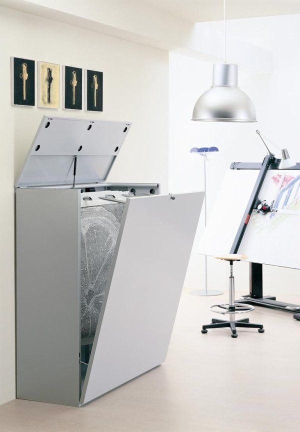 In uno studio, accanto a un tavolo da disegno, una sedia e una lampada a sospensione, si trova un armadio verticale aperto di colore grigio per documenti.