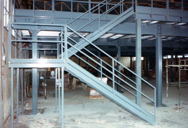 All'interno di un magazzino, una scala industriale in metallo grigio con pianerottolo intermedio e ringhiera conduce a una piattaforma sopraelevata.