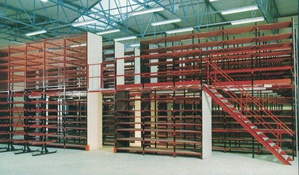 Sistema di scaffalature multilivello per magazzino con montanti in acciaio rosso, ripiani neri e una scala rossa che conduce al livello superiore.