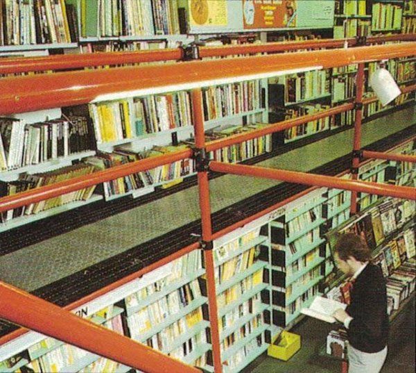 Una persona legge un libro in una biblioteca con scaffalature a più livelli collegate da impalcature metalliche arancioni.