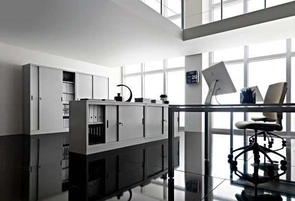 Ufficio moderno con armadietti bianchi, scrivania in vetro, computer e sedia su un pavimento lucido e riflettente.