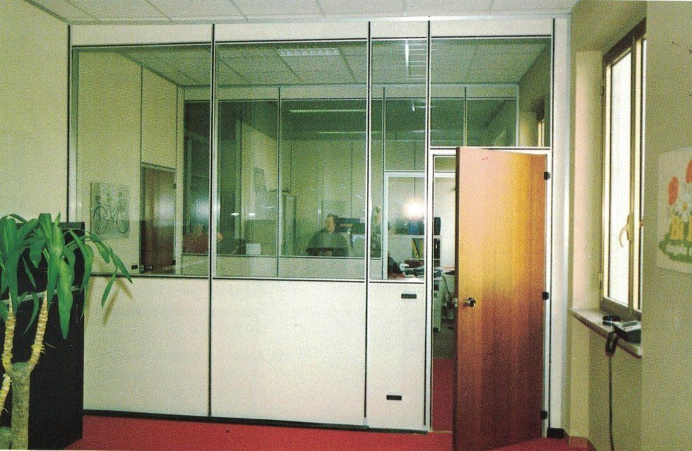 Una parete divisoria modulare in vetro e legno per ufficio, in una stanza con pavimento in moquette rossa e porta in legno.