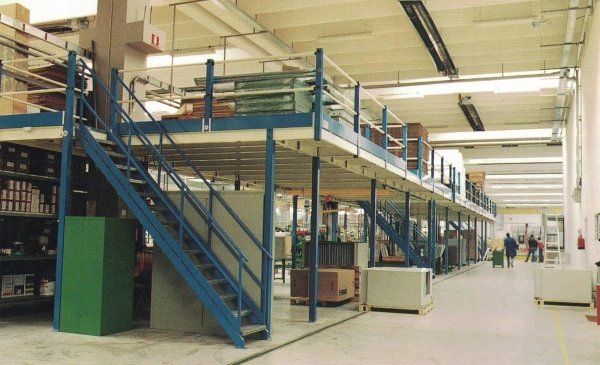 Struttura a soppalco industriale blu con scala all'interno di un magazzino, con operai visibili in lontananza.