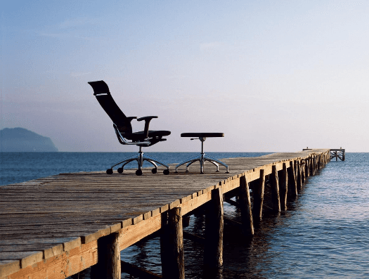 Una sedia da ufficio ergonomica e un piccolo sgabello sono posizionati su un pontile di legno che si protende nelle calme acque blu dell'oceano.