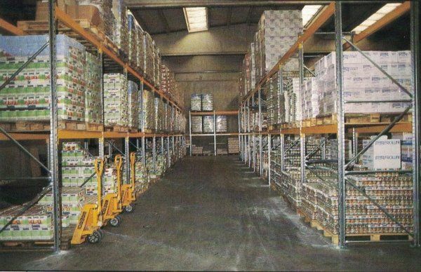 Un corridoio di magazzino caratterizzato da alte scaffalature metalliche piene di scatole pallettizzate, con transpallet gialli sul pavimento.