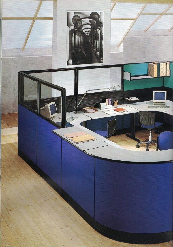 Una moderna postazione di lavoro in ufficio, dalle forme curve e di colore blu, con computer, lampada da scrivania e scaffali, su un pavimento in legno chiaro.