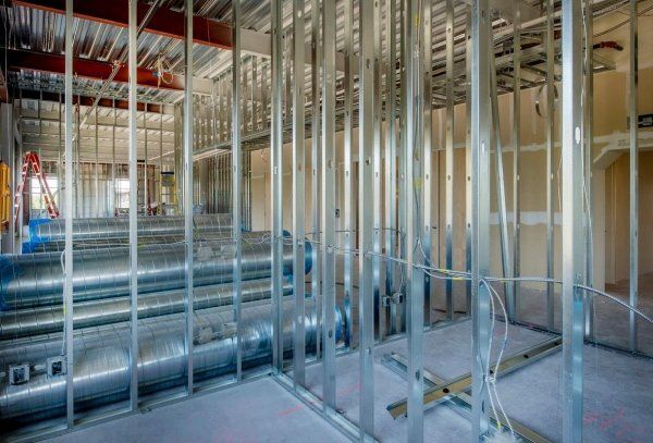 Un cantiere edile interno che mostra la struttura metallica delle pareti, le travi del soffitto a vista e i grandi condotti di ventilazione cilindrici.