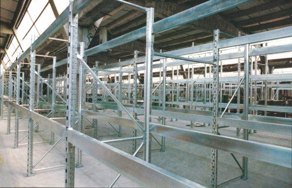 In un magazzino con soffitto alto, si trovano scaffalature industriali metalliche vuote con telai a traversa.
