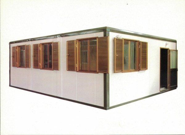 Un edificio modulare prefabbricato di colore bianco, con persiane in legno alle finestre e una porta aperta sul lato destro.
