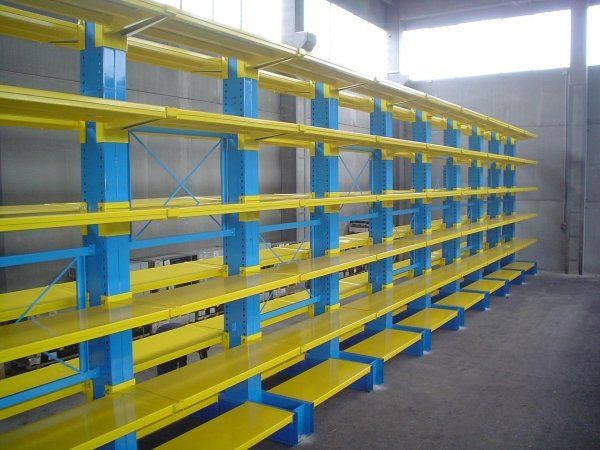 In un magazzino, una lunga fila di scaffalature industriali vuote con pilastri di supporto blu e ripiani orizzontali gialli.