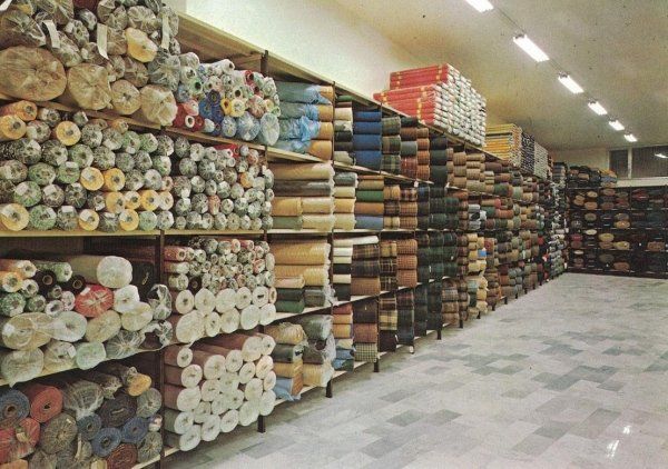 L'interno di un magazzino di un negozio di tessuti, con file di alti scaffali metallici pieni di numerose e colorate pezze di tessuto arrotolato.