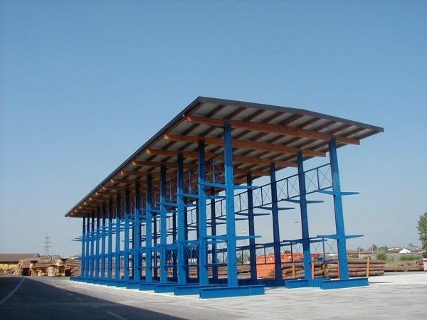 Una struttura di stoccaggio a sbalzo in metallo blu, alta e con tetto, si erge in un cortile esterno pavimentato sotto un cielo azzurro e limpido.