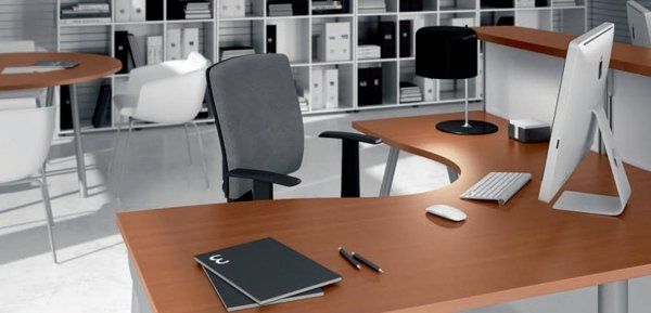 Postazione di lavoro moderna per ufficio, con scrivania curva in legno, sedia grigia, computer e scaffalature in un ambiente minimalista.