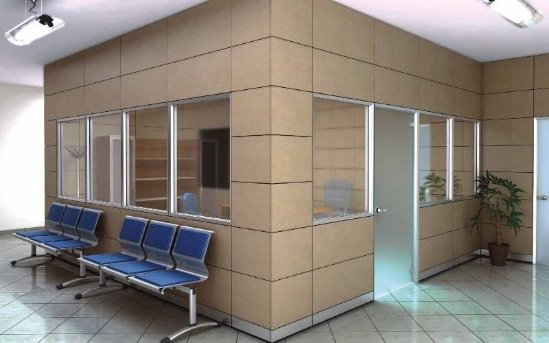 Una sala d'attesa con file di sedie blu di fronte a un ufficio con pareti in vetro, pannelli color beige e pavimento piastrellato.