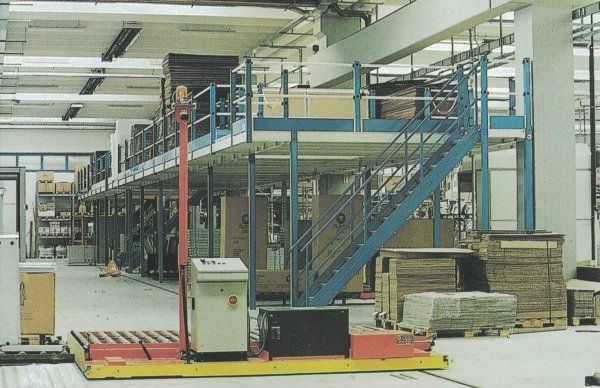 Un carrello elevatore automatico giallo per pallet opera all'interno di un magazzino dotato di un soppalco industriale a più livelli e di scale.