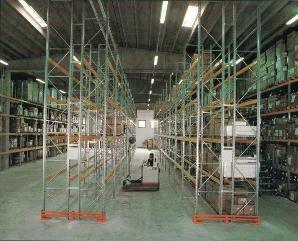 Interno di un magazzino caratterizzato da file di scaffalature metalliche industriali cariche di merci, illuminate da faretti a soffitto.