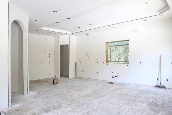 Una stanza non rifinita con pareti in cartongesso bianco, soffitto ribassato, una finestra, una porta ad arco e una scopa appoggiata a una parete.