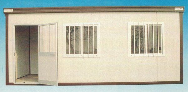 Una cabina modulare portatile con una sola porta, due finestre con telaio bianco e inferriate di sicurezza, e pareti esterne color bianco sporco.